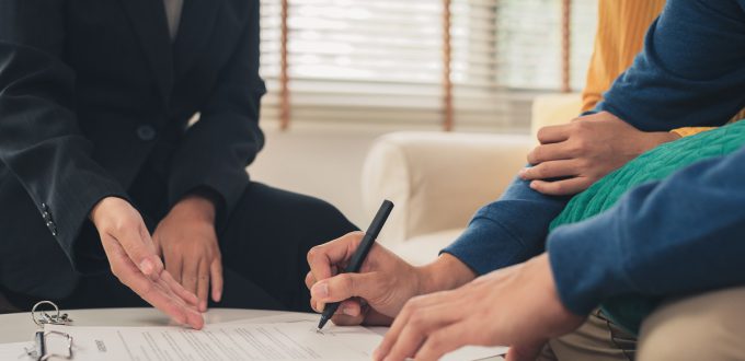 Happy young Asian couple and realtor agent. Cheerful young man signing some documents while sitting at desk together with his wife. Buying new house real estate. Signing good condition contract.