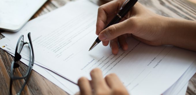 Unrecognizable businessman examining papers at table. Manager with ballpoint pen filling business papers. Close-up of male hands working at desk. Analyzing documents concept